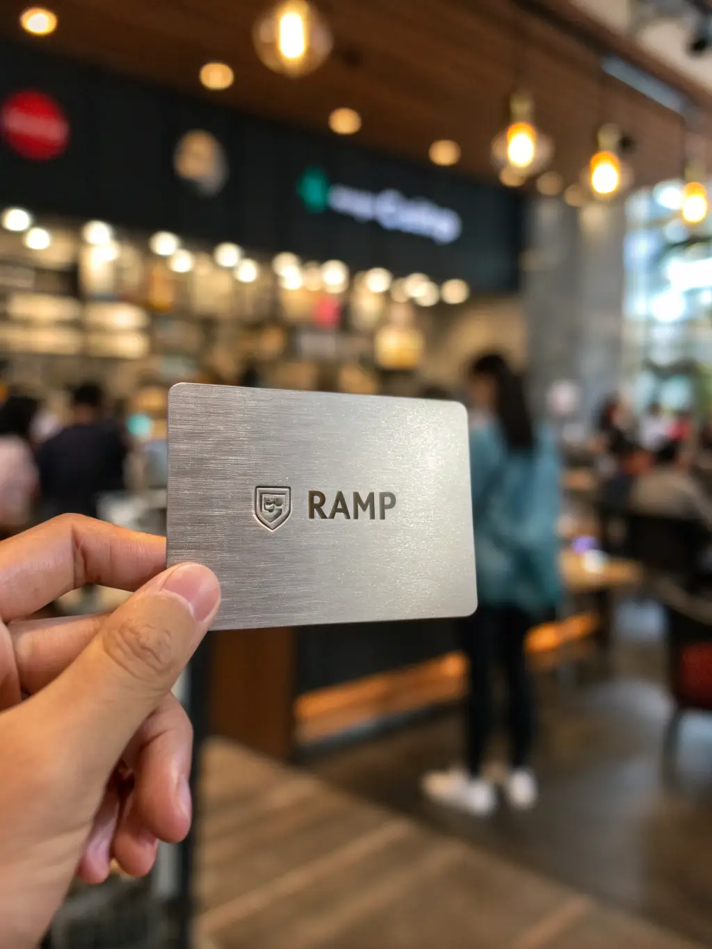 A close-up shot of the Ramp card, emphasizing its metallic finish and the Ramp logo. The background is a blurred image of a busy entrepreneur working at a coffee shop.